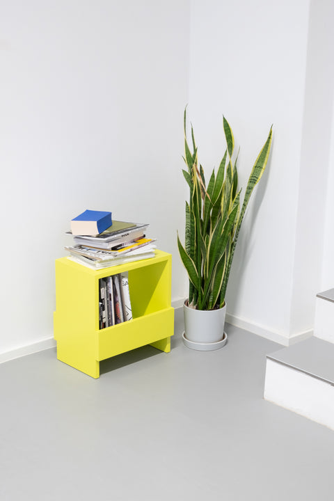 Ein Hocker oder Beistelltisch aus Massivholz mit Stauraum in schwefelgelb gefüllt mit Büchern und Zeitschriften und einer Pflanze daneben